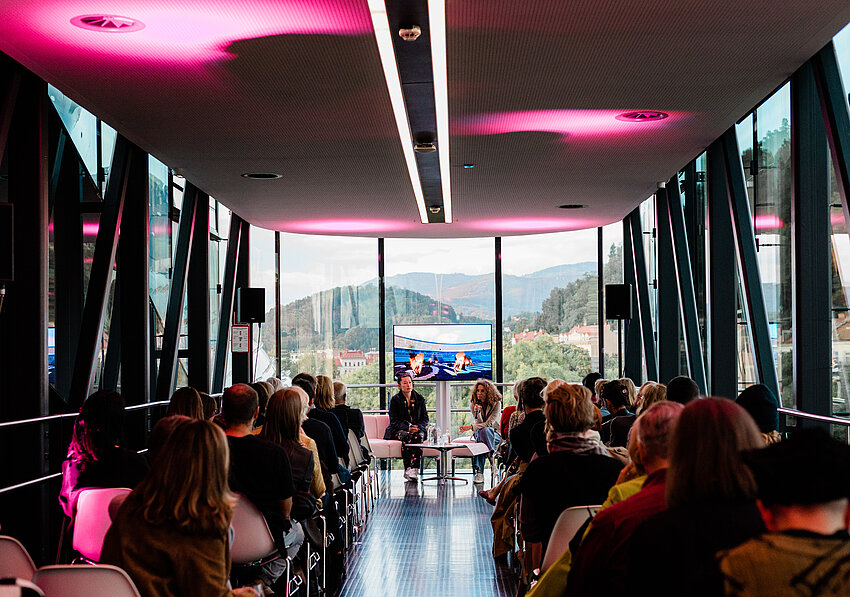 A group of people are lined up in the Needle at Kunsthaus Graz, seen from behind. At the end of the Needle, facing the camera, sit artist Hito Steyerl and curator Katrin Bucher Trantow in conversation, behind them a screen showing excerpts from a film. Through the glass façade of the Needle, the green hilly landscape of Graz can be seen outside.