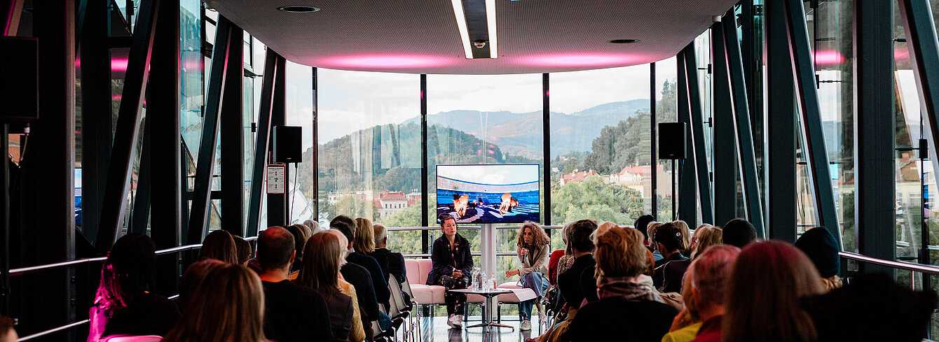  Eine Gruppe an Menschen sitzen aufgereiht in der Needle des Kunsthauses Graz, man sieht sie von hinten. Am Ende der Needle, der Kamera zugewandt, sitzen Künstlerin Hito Steyerl und Kuratorin Katrin Bucher Trantow im Gespräch, hinter ihnen ein Bildschirm der Ausschnitte aus einem Film zeigt. Durch die Glasfassade der Needle erkennt man draußen die grüne Hügellandschaft von Graz. 