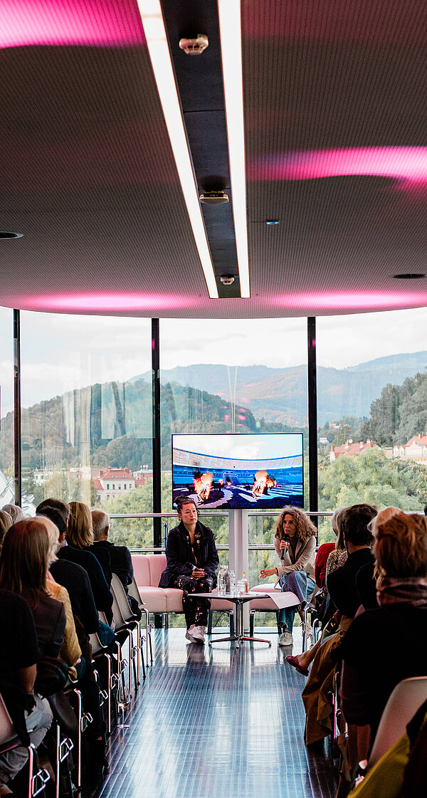 A group of people are lined up in the Needle at Kunsthaus Graz, seen from behind. At the end of the Needle, facing the camera, sit artist Hito Steyerl and curator Katrin Bucher Trantow in conversation, behind them a screen showing excerpts from a film. Through the glass façade of the Needle, the green hilly landscape of Graz can be seen outside.