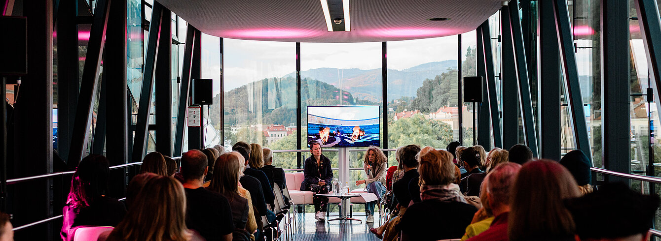A group of people are lined up in the Needle at Kunsthaus Graz, seen from behind. At the end of the Needle, facing the camera, sit artist Hito Steyerl and curator Katrin Bucher Trantow in conversation, behind them a screen showing excerpts from a film. Through the glass façade of the Needle, the green hilly landscape of Graz can be seen outside.
