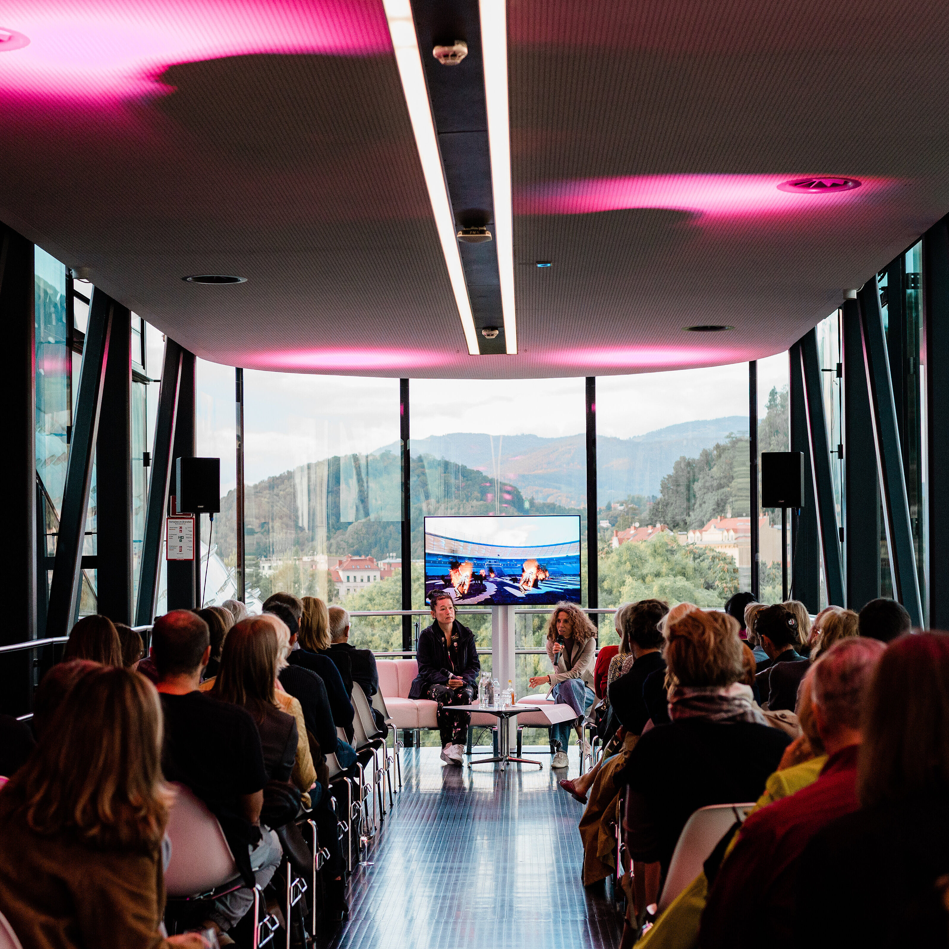 Eine Gruppe an Menschen sitzen aufgereiht in der Needle des Kunsthauses Graz, man sieht sie von hinten. Am Ende der Needle, der Kamera zugewandt, sitzen Künstlerin Hito Steyerl und Kuratorin Katrin Bucher Trantow im Gespräch, hinter ihnen ein Bildschirm der Ausschnitte aus einem Film zeigt. Durch die Glasfassade der Needle erkennt man draußen die grüne Hügellandschaft von Graz. 