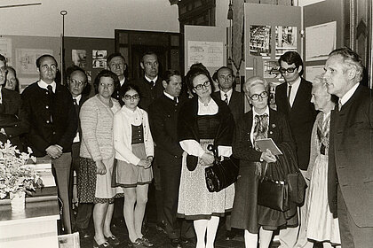 Schwarz-weiß Fotografie von einer Gruppe von Menschen. Diese stehen in einem Ausstellungsraum und sehen einem Mann zu, der zur Gruppe spricht.