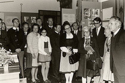 Black and white photography of a group of people. They stand in an exhibition room and watch a man who speaks to the group.