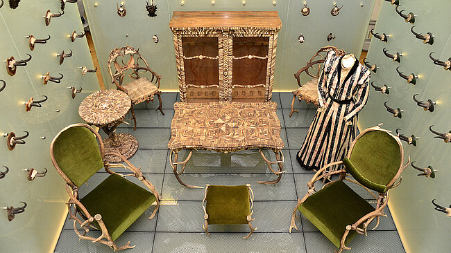 In a showcase are several pieces of furniture and a mannequin wearing a striped dress. The furniture is covered with green velvet and is otherwise made entirely of antler and horn.