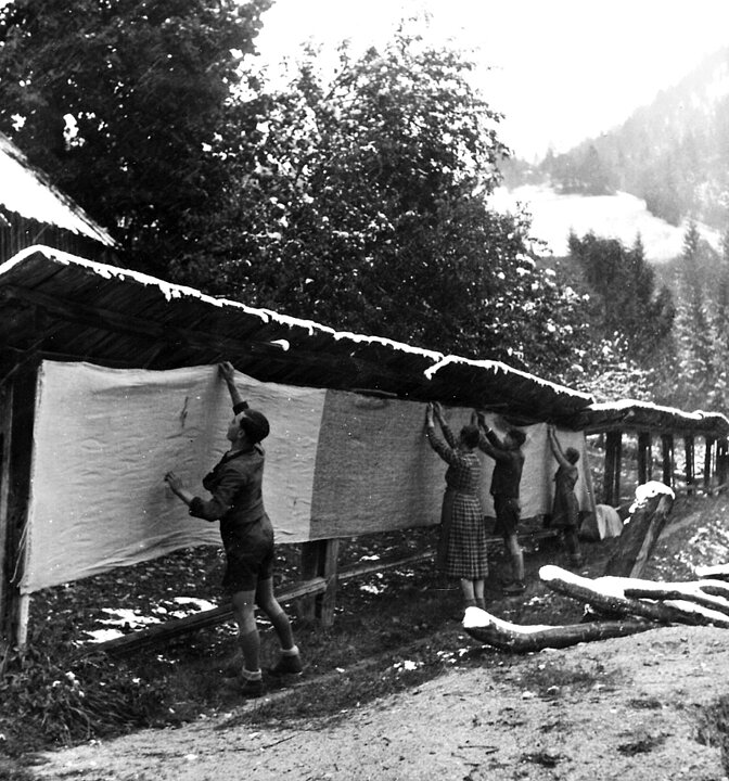 Alte schwarz-weiß Fotografie von einer Gruppe von Männern, die eine lange Stoffbahn unter einem schmalen Dach aufhängt. 