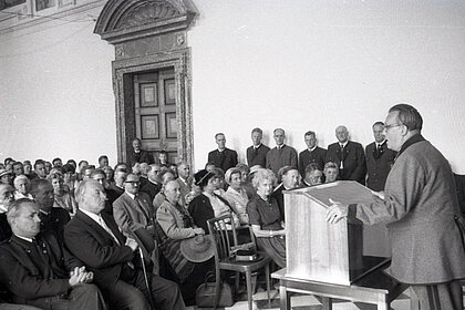 Eine Schwarz-weiß Fotografie. Rechts im Bild steht ein Mann, der eine Rede hält. Links sitzen und stehen Erwachsene und hören dem Redner zu. Das Publikum trägt teilweise Tracht.