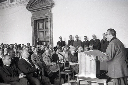 
A black and white photograph. On the right of the picture there is a man giving a speech. On the left, adults sit and stand and listen to the speaker. Some of the audience wear traditional costumes.