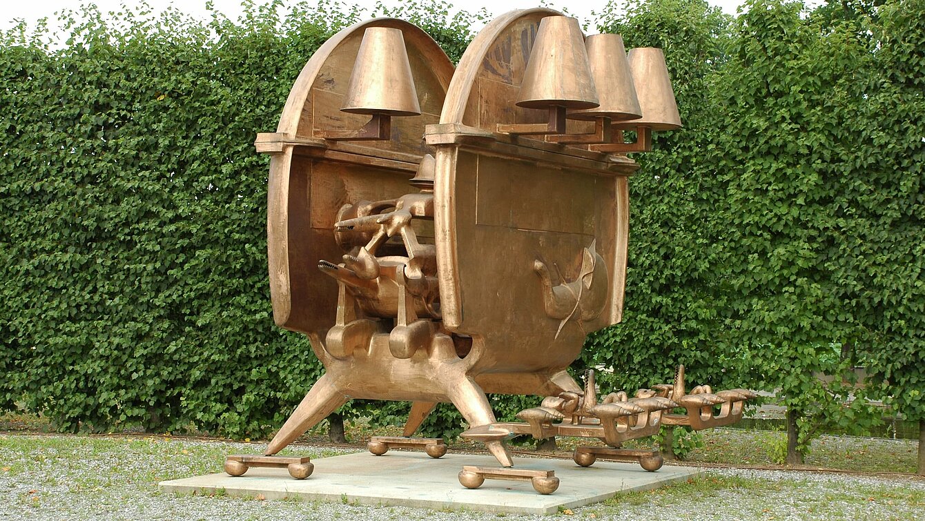 Die Skulptur ist übergroß, kupferfarben und steht auf einem flachen Sockel. Sie steht auf vier Füßen und hat mechanische und und biologische Elemente, die diesem Objekt etwas Lebendiges verleiht.  