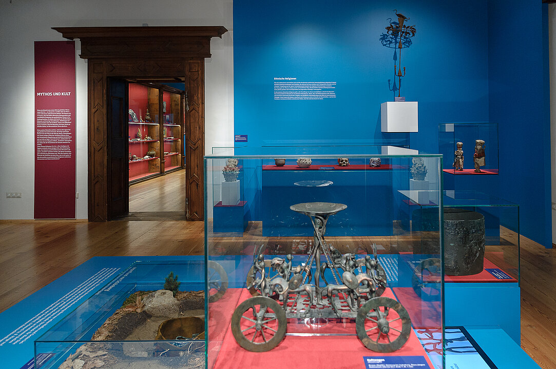 In einem Raum mit blauen Wänden stehen zahlreiche Vitrinen, in diesen sind archäologische Objekte zu sehen.