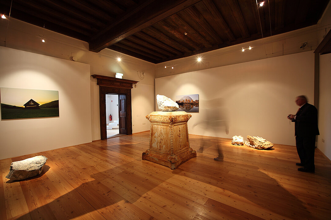 Ausstellungsansicht von einem Raum. In der Mitte des Raumes steht eine antikwirkender Pfeiler, auf dem ein Stein liegt. Im restlichen Raum liegen größere Steine als Kunstinstallationen verteilt.