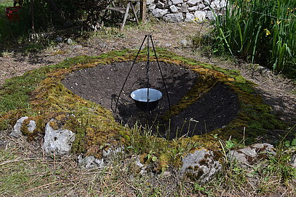 Am Foto ist ein Garten mit Gartenstuhl, Steinmauer, Pflanzen, Bäumen. Im Hintergrund sieht man zwei Gebäude. Im Vordergrund ist ein Loch in der Erde, in dem in der Mitte ein Kessel auf einem Dreibein hängt. Rund um das Loch liegt Moos.