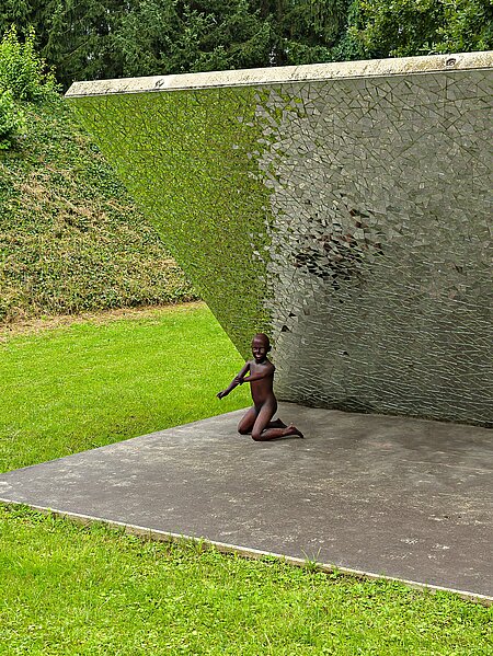 Im Berggarten des Skulpturenparks steht eine diagonal aufragende Betonwand. Sie ist unterseits mit kleinen Spiegelstücke versehen. Die Umwelt wird durch das Sonnenlicht in den Scherben gebrochen. 