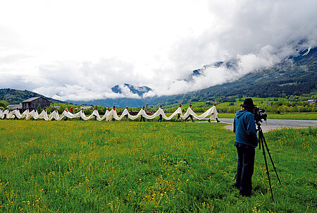Photography of a row of men walking through a foggy mountain landscape. The men are connected by a long white ribbon. On the right front of the picture stands a man with a film camera
