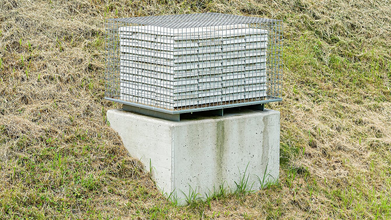 Die Skulptur verwehrt den Zugang zu 999 Barren aus feinstem Beton. Die fein säuberlich gestapelten Barren sind von einem Gitter umgeben. 