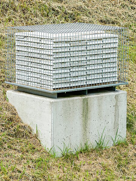 Die Skulptur verwehrt den Zugang zu 999 Barren aus feinstem Beton. Die fein säuberlich gestapelten Barren sind von einem Gitter umgeben. 