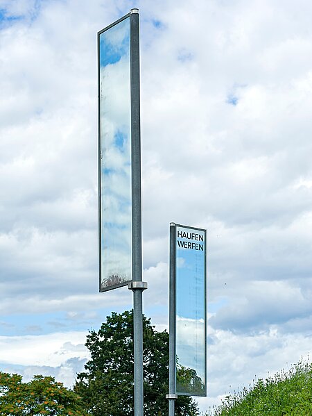 Two mirrors on poles stand on a hill between two paths. As they constantly turn in the wind, they constantly reflect different things. What is reflected in them symbolizes a view into two different worlds. For the moments in which the mirrors are reflected in each other, a connection is created between these two worlds.