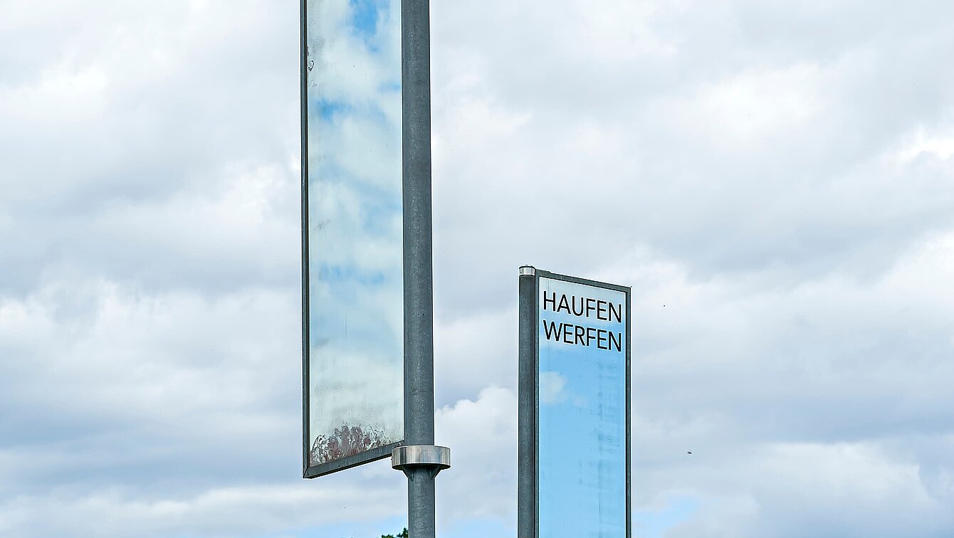 Auf einem Hügel zwischen zwei Wegen stehen zwei Spiegel auf Stangen. Durch ihre ständige Drehung im Wind geben sie stetig anderes wieder. Das sich in ihnen Spiegelnde symbolisiert einen Blick in zwei unterschiedliche Welten. Für die Momente, in denen sich die Spiegel ineinander spiegeln, entsteht eine Verbindung dieser beiden Welten.