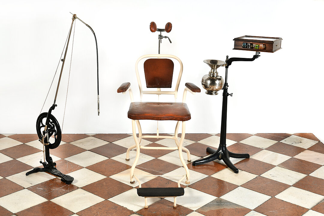  A historical dentist chair made of white metal and brown leather. To the left of it is a drill and to the right of it is a metal spittoon