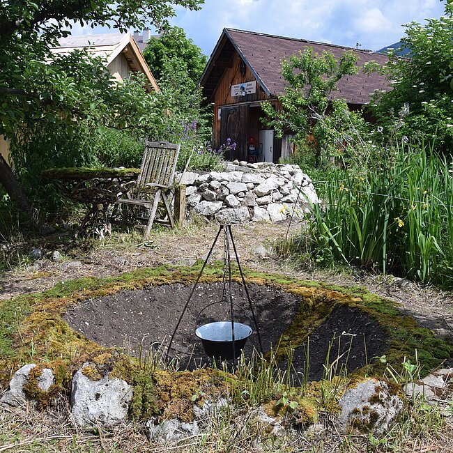 Am Foto ist ein Garten mit Gartenstuhl, Steinmauer, Pflanzen, Bäumen. Im Hintergrund sieht man zwei Gebäude. Im Vordergrund ist ein Loch in der Erde, in dem in der Mitte ein Kessel auf einem Dreibein hängt. Rund um das Loch liegt Moos.