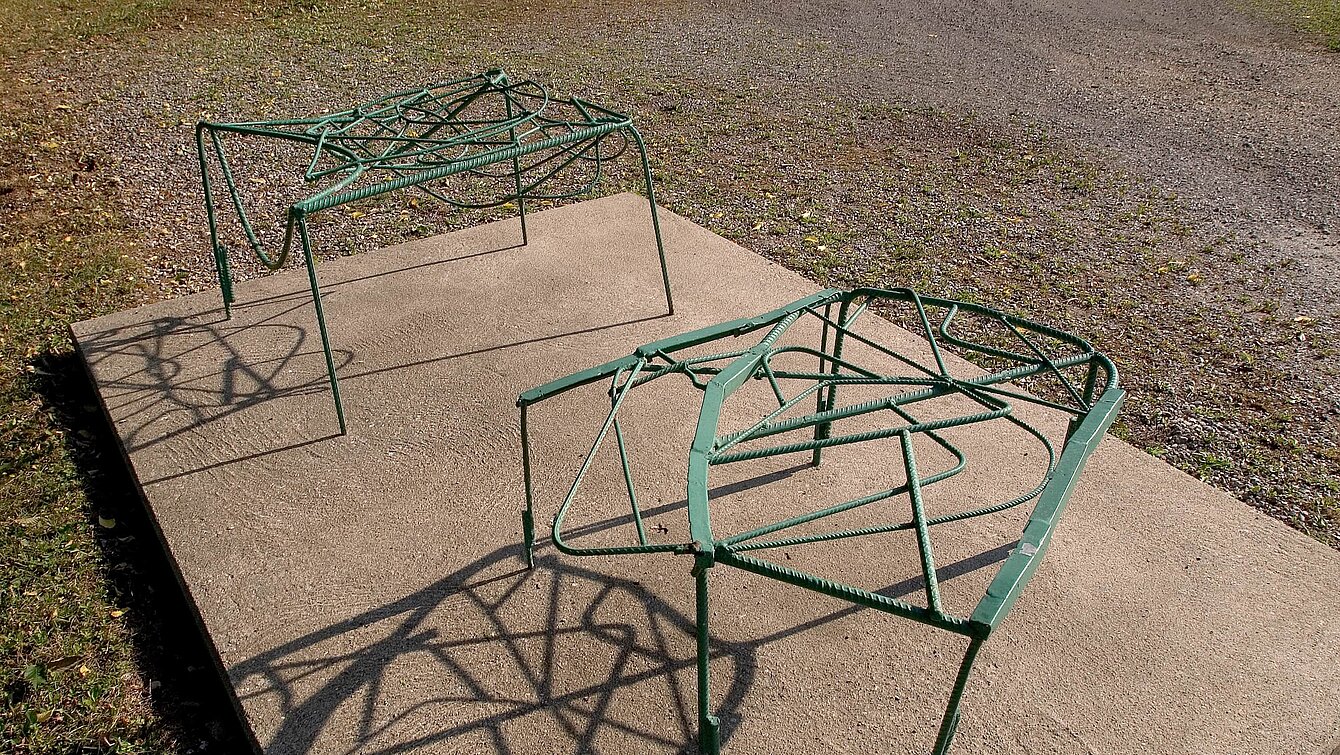 The two-part sculpture made of bent and green-painted metal struts resembles two stools in shape and rests on a concrete base.