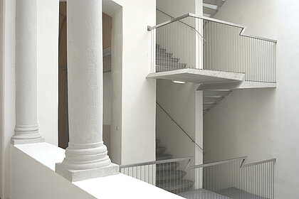 Weißes Treppenhaus mit metallenem Geländer. Im Vordergrund stehen zwei Säulen