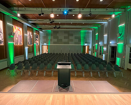 View from the stage in the Heimatsaal in the Folk Life Museum. In the foreground is a lectern, in the background are chairs arranged in rows. Pictures hang on the walls and the room is illuminated in color.