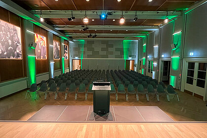 View from the stage in the Heimatsaal in the Folk Life Museum. In the foreground is a lectern, in the background are chairs arranged in rows. Pictures hang on the walls and the room is illuminated in color.