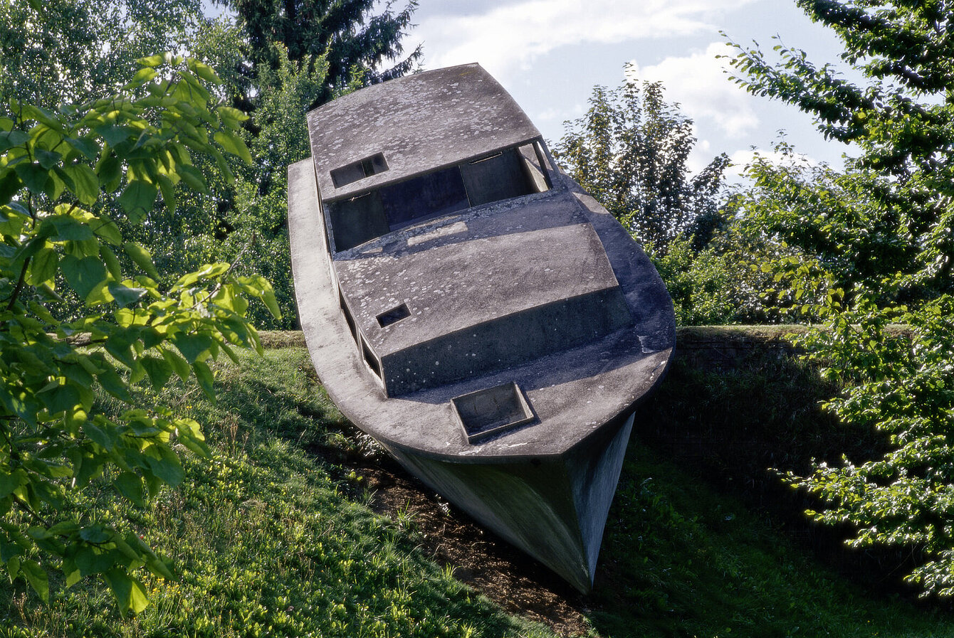 Eine Betonskulptur in der Form eines realen Bootes scheint auf der Hügellandschaft des Skulpturenparks zu schwimmen. 