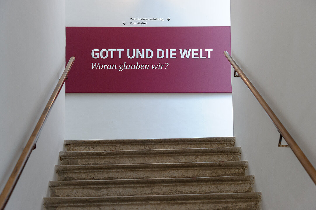 Foto von einem Treppenaufgang. Am Ende der Stiege ist der Schriftzug "Gott und die Welt" vor einer roten Wand zu sehen.
