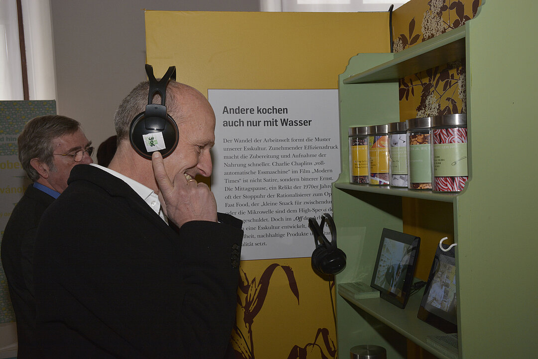 Ein Mann mit großen Kopfhörern steht vor einen hellgrünen Ausstellungsregal und schaut auf einem Bildschirm ein Video.