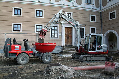 An excavator is standing in front of the castle entrance and pipes are being laid.