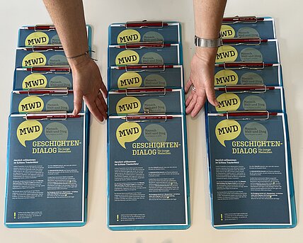 Drei Reihen Clipboards mit dem ausgedruckten Fragebogen "Geschichten_Dialog". Zwei Hände ordnen die Clipboards.