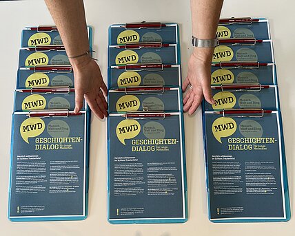 Drei Reihen Clipboards mit dem ausgedruckten Fragebogen "Geschichten_Dialog". Zwei Hände ordnen die Clipboards.