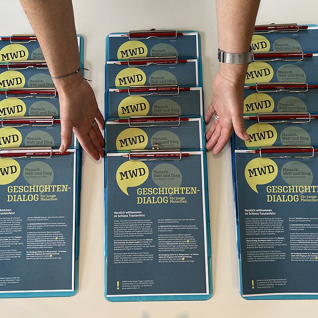 Drei Reihen Clipboards mit dem ausgedruckten Fragebogen "Geschichten_Dialog". Zwei Hände ordnen die Clipboards.
