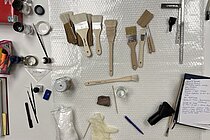 Auf einer Tischplatte ist eine Vielzahl an Utensilien wie ein Pinsel, ein Glas mit einer Flüssigkeit zu sehen, die in der Präventiven Konservierung verwendet werden.