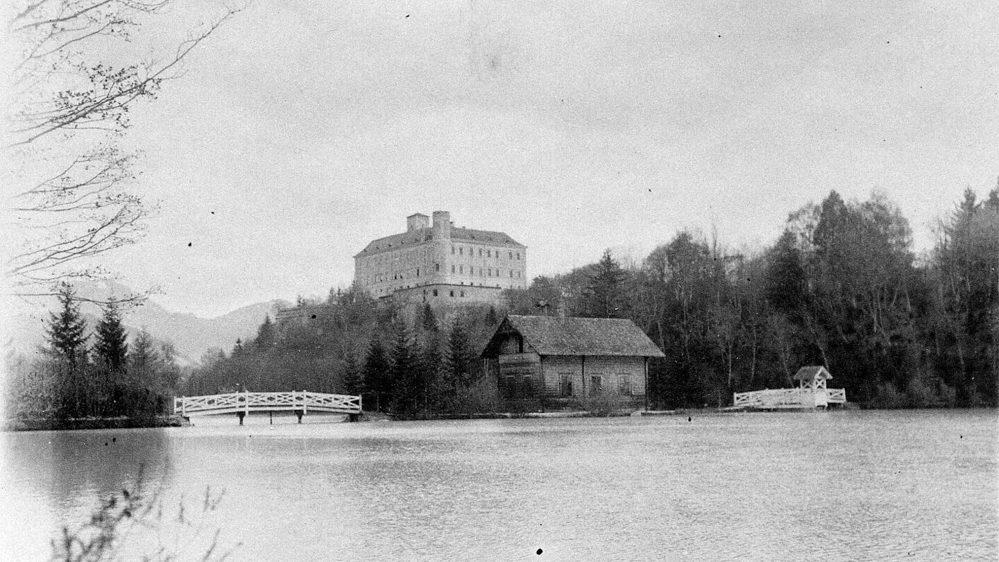 
Black and white photography of Trautenfels Castle can be seen. There is a lake in the foreground. The sky is clouded over.