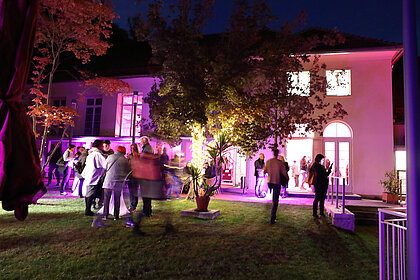 Menschen stehen im beleuchteten Duftgarten bei einer Abendveranstaltung im Heimatsaal.