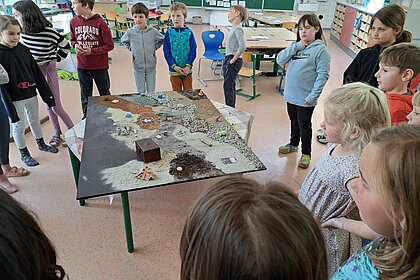 Zu sehen ist eine Gruppe von Kindern, die um einen Tisch stehen, auf dem unterschiedliche Gegenstände zu sehen sind.