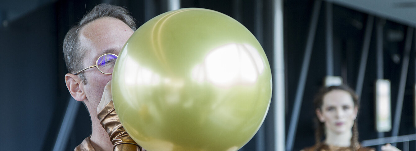 Footage of a performance in the Needle at the Kunsthaus Graz. In an extreme close-up and cropped shot, a man blows up a golden balloon. Another performer can be seen out of focus in the background.