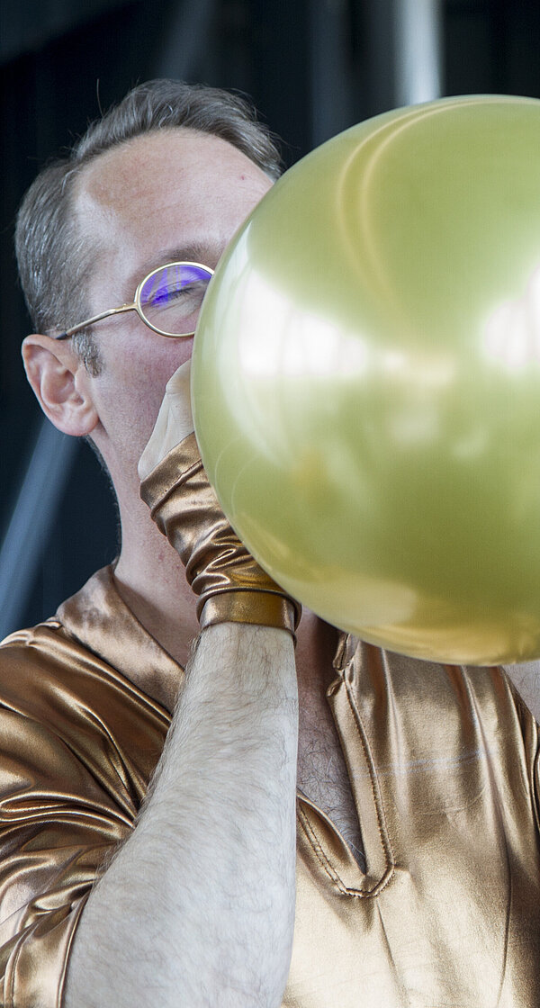 Footage of a performance in the Needle at the Kunsthaus Graz. In an extreme close-up and cropped shot, a man blows up a golden balloon. Another performer can be seen out of focus in the background.
