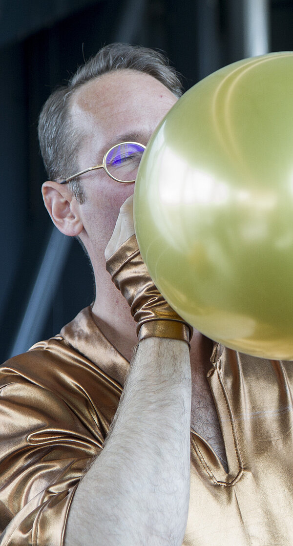 Aufnahmen einer Performance in der Needle des Kunsthauses Graz. In einer extremen Nahaufnahme und angeschnitten bläst ein Mann einen goldenen Luftballon auf. Unscharf im Hintergrund ist eine weitere Performerin zu sehen.