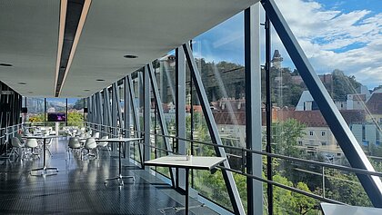 In der Needle im Kunsthaus Graz stehen im Vordergrund Stehtische. Im Hintergrund sind kleine Kaffeehaus-Sitzgruppen aufgestellt.