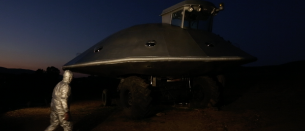 Person in a silver full-body suit walks towards a UFO hovering in the sky at dusk.