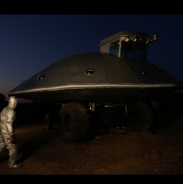 Person in a silver full-body suit walks towards a UFO hovering in the sky at dusk.