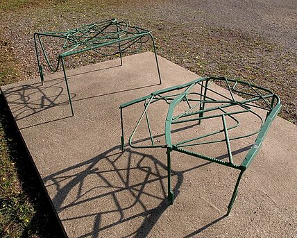 Die zweiteilige Skulptur aus gebogenen und grün lackierten Metallstreben ähnelt in ihrer Form zwei Hockern und ruht auf einem Betonsockel.