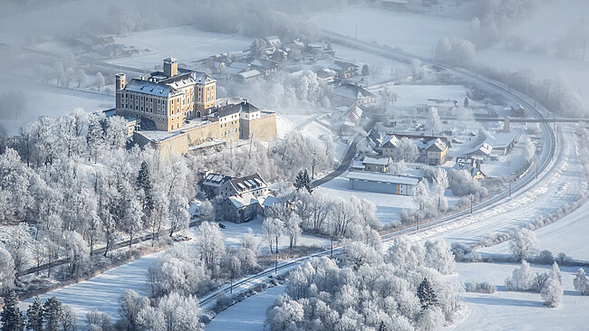 Schloss Trautenfels aus der Luft fotografiert. Überall liegt Schnee. Vor dem Schloss laufen Bahngleise entlang. An der Bahnstrecke entlang stehen Häuser und Bäume.