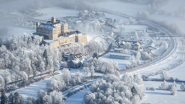 Trautenfels Castle photographed from the air. There is snow everywhere. Railroad tracks run along in front of the castle. Along the railroad line there are houses and trees.