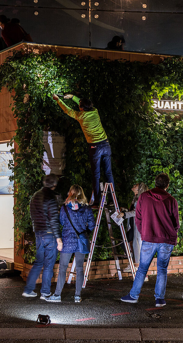 People gather in front of the Kunsthaus Graz at night. On the forecourt is a small building overgrown with hop plants. The artist Alfredo Barsuglia stands on a ladder and harvests the hops.