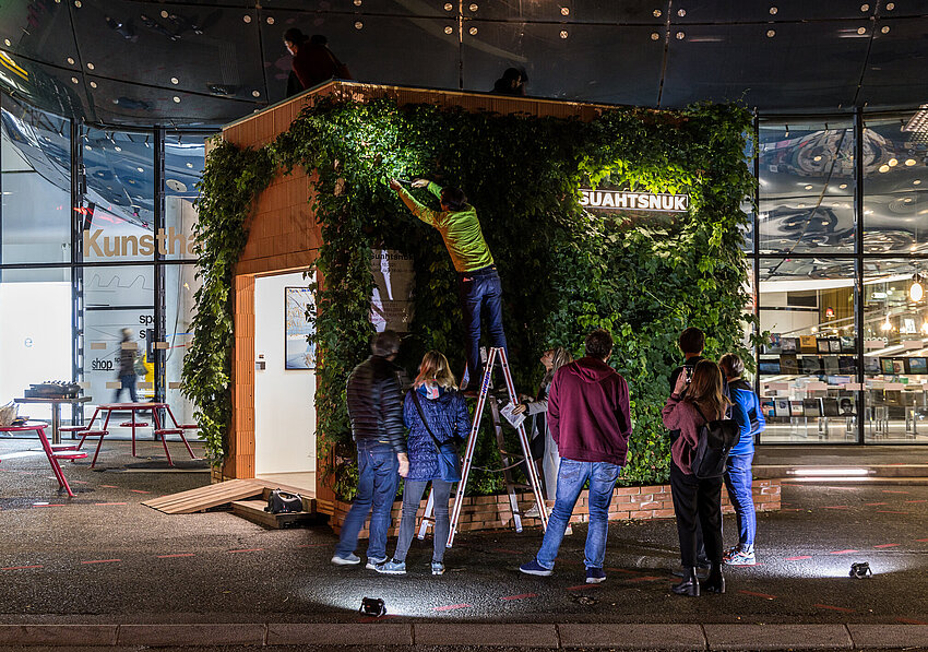 People gather in front of the Kunsthaus Graz at night. On the forecourt is a small building overgrown with hop plants. The artist Alfredo Barsuglia stands on a ladder and harvests the hops.