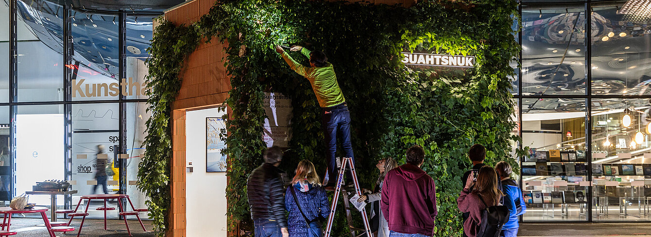 People gather in front of the Kunsthaus Graz at night. On the forecourt is a small building overgrown with hop plants. The artist Alfredo Barsuglia stands on a ladder and harvests the hops.